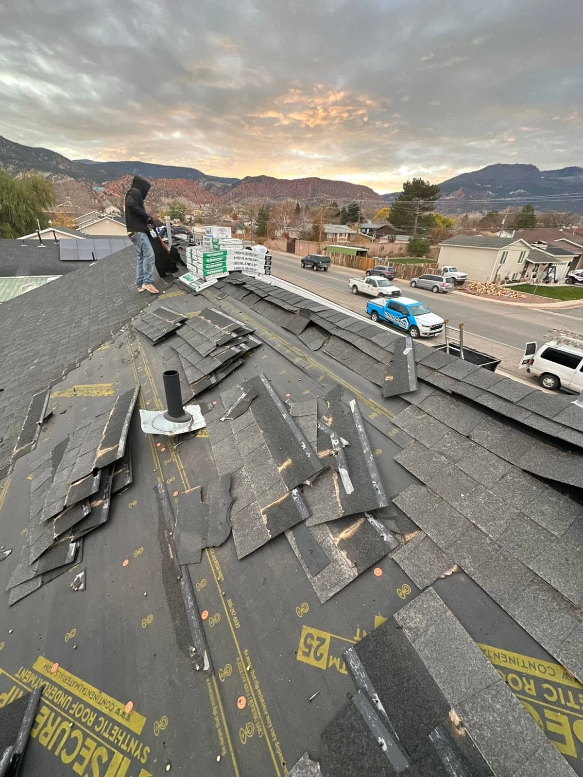Roof replacement underway at dusk with new shingle bundles staged and red-rock hills behind